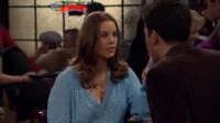 Mulher de cabelo castanho em blusa azul conversa com homem de costas em bar. Pessoas borradas ao fundo, clima social.