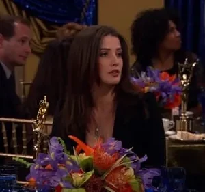 Mulher morena com expressão atenta em jantar de gala. Há um arranjo floral e uma estatueta dourada na mesa. Outras pessoas ao fundo.