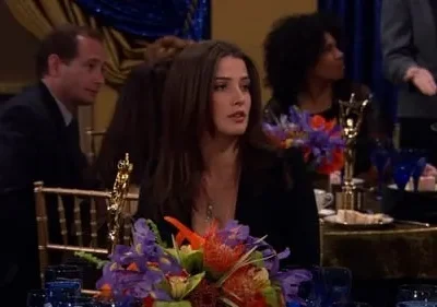 Mulher morena com expressão atenta em jantar de gala. Há um arranjo floral e uma estatueta dourada na mesa. Outras pessoas ao fundo.