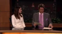 Dois apresentadores de noticiário sentados à bancada em estúdio com skyline noturno. Mulher escuta, homem manuseia papéis. Clima formal.