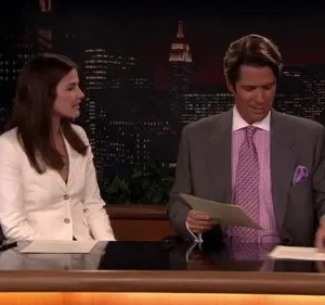 Dois apresentadores de noticiário sentados à bancada em estúdio com skyline noturno. Mulher escuta, homem manuseia papéis. Clima formal.