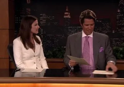 Dois apresentadores de noticiário sentados à bancada em estúdio com skyline noturno. Mulher escuta, homem manuseia papéis. Clima formal.