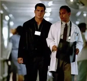 Dois homens sérios caminham em corredor de hospital. Um médico com jaleco branco segura exames. O outro, de jaqueta preta e crachá.