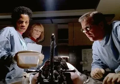 Três investigadores forenses em scrubs examinam um corpo em uma mesa de autópsia. Uma mulher segura um recipiente, um homem de óculos observa. Cena séria.