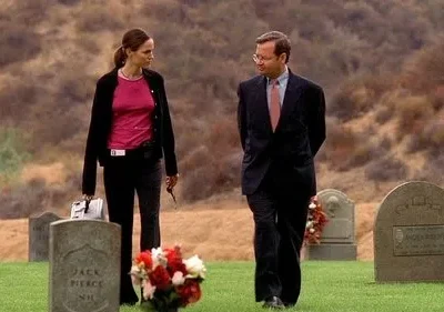 Mulher de blusa rosa e homem de terno caminham sérios em um cemitério com colinas ao fundo. Ambiente sombrio.