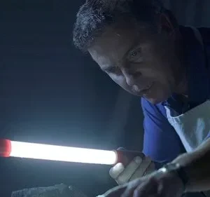 Homem de camisa azul e avental branco foca uma luz brilhante de lanterna em um objeto, imerso em escuridão. Atmosfera de investigação.