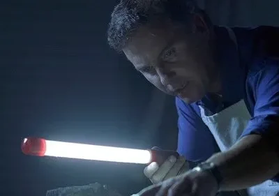 Homem de camisa azul e avental branco foca uma luz brilhante de lanterna em um objeto, imerso em escuridão. Atmosfera de investigação.