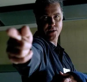 Homem sério em uniforme escuro aponta diretamente para o espectador. Ambiente institucional com um relógio na parede.