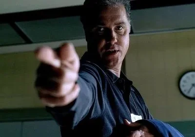 Homem sério em uniforme escuro aponta diretamente para o espectador. Ambiente institucional com um relógio na parede.