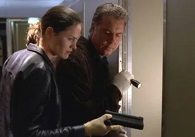 Dupla em luvas brancas investiga um local escuro. Homem ilumina com lanterna, mulher segura equipamento. Clima de mistério.