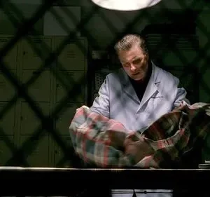 Homem de jaleco branco examina camisa xadrez sobre mesa. Cenário escuro com sombras em grade e armários ao fundo.