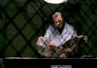 Homem de jaleco branco examina camisa xadrez sobre mesa. Cenário escuro com sombras em grade e armários ao fundo.