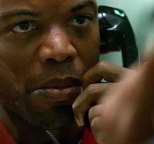 Homem negro em uniforme vermelho, close, com expressão intensa ao falar ao telefone preto. Fundo borrado, clima tenso.