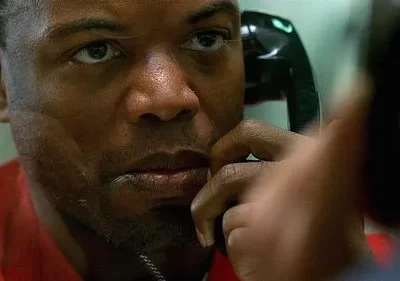 Homem negro em uniforme vermelho, close, com expressão intensa ao falar ao telefone preto. Fundo borrado, clima tenso.