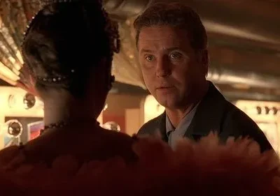 Homem sério de jaqueta olha para frente, com mulher de costas em traje elaborado e penas no primeiro plano. Cenário de show noturno.