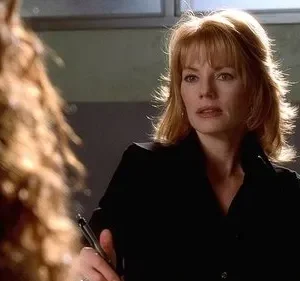 Mulher de cabelo ruivo e franja, camisa escura, olha intensamente para pessoa desfocada. Cenário interno, clima sério.