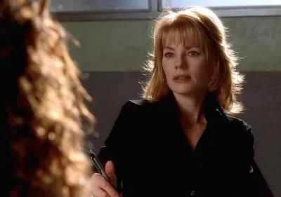 Mulher de cabelo ruivo e franja, camisa escura, olha intensamente para pessoa desfocada. Cenário interno, clima sério.