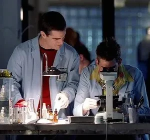 Dois homens em jalecos trabalham em um laboratório. Um manipula tubos de ensaio, o outro usa um microscópio. Ambiente focado em pesquisa.
