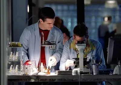 Dois homens em jalecos trabalham em um laboratório. Um manipula tubos de ensaio, o outro usa um microscópio. Ambiente focado em pesquisa.