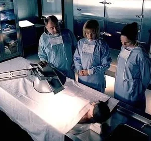 Num necrotério, três legistas em scrubs e máscaras observam atentamente um corpo numa mesa de autópsia iluminada. Clima sério e investigativo.