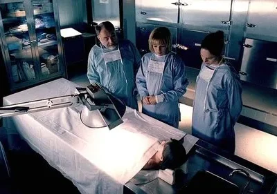 Num necrotério, três legistas em scrubs e máscaras observam atentamente um corpo numa mesa de autópsia iluminada. Clima sério e investigativo.