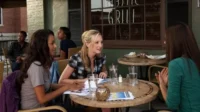 Três mulheres conversam em mesas de um café ao ar livre. Uma loira sorri para a morena. Outros clientes no fundo. Cena casual.