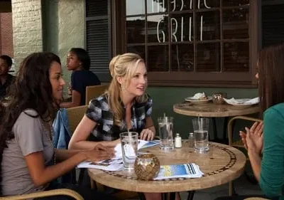 Três mulheres conversam em mesas de um café ao ar livre. Uma loira sorri para a morena. Outros clientes no fundo. Cena casual.