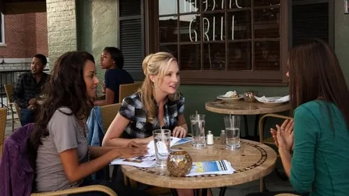 Três mulheres conversam em mesas de um café ao ar livre. Uma loira sorri para a morena. Outros clientes no fundo. Cena casual.
