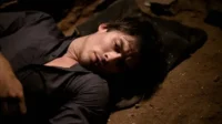 Diários de um Vampiro – T01E05:Você Esta Morto Para Mim 7 Homem de camisa escura deitado no chão de terra,com olhos semiabertos. Parece inconsciente ou exausto num ambiente escuro e tenso.