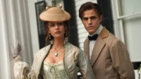 Diários de um Vampiro – T01E06:Garotas Perdidas 6 Mulher de chapéu e vestido de época e homem de terno sérios em frente a uma mansão colonial. Ambiente de drama e mistério.