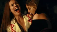 Diários de um Vampiro – T01E07:Assombrada 5 Mulher de presas morde pescoço de outra que grita. Cena noturna intensa com clima de horror e dor.