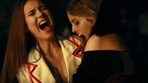 Mulher de presas morde pescoço de outra que grita. Cena noturna intensa com clima de horror e dor.