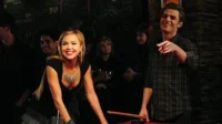 Diários de um Vampiro – T01E08:162 Velas 4 Mulher loira sorridente e homem de xadrez com taco de sinuca num bar escuro. Ambos felizes num ambiente jovem e social.
