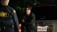 Diários de um Vampiro – T01E10:A Descoberta 3 Um xerife de costas confronta um jovem de jaqueta de couro com expressão assustada,ao lado de um SUV à noite. Cena tensa.