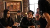 Diários de um Vampiro – T01E11:Ascendência 2 Mulher séria e homem de jaqueta de couro em bar rústico,olhando uma mulher de costas. Pôster de cerveja. Clima intrigante.