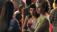 Diários de um Vampiro – T01E12:Visita Desagradável 1 Três mulheres numa festa. Uma de costas,uma loira de cardigan e uma morena com coquetel,ambas sérias. Fundo festivo,balões.