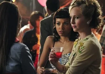 Três mulheres numa festa. Uma de costas, uma loira de cardigan e uma morena com coquetel, ambas sérias. Fundo festivo, balões.