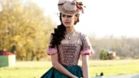 Mulher com vestido histórico rosa xadrez e saia verde,chapéu de palha,em campo ensolarado. Ela tem olhar pensativo.