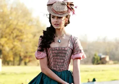 Mulher com vestido histórico rosa xadrez e saia verde, chapéu de palha, em campo ensolarado. Ela tem olhar pensativo.
