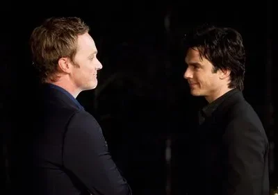 Dois homens com cabelos claros e escuros se encaram, sorrindo gentilmente em um fundo escuro.