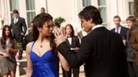 Mulher em vestido azul e homem de terno se encaram intensamente,ele estende a mão. Fundo de evento formal.