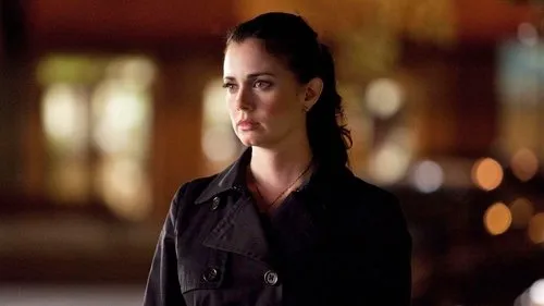Diários de um Vampiro – T01E21:Isobel 1 Mulher séria de cabelo escuro preso e casaco preto olha para o lado. Fundo noturno com luzes desfocadas cria clima sombrio.