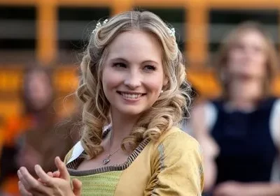 Mulher loira sorridente com vestido medieval dourado e verde, cabelo cacheado e tiara, aplaude. Fundo com ônibus escolar.