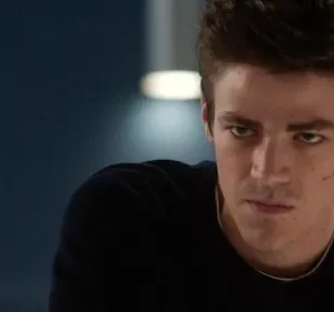 Um jovem de cabelo escuro, com cicatriz na bochecha, olha intensamente para frente. Expressão séria, em um ambiente escuro e dramático.