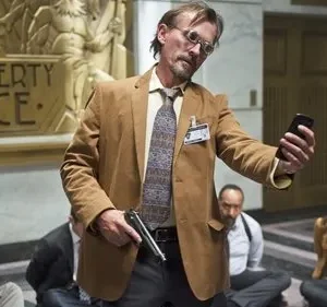 Um homem com arma e óculos filma reféns amarrados no chão de um prédio com JUSTICE no fundo. Cena tensa de sequestro.