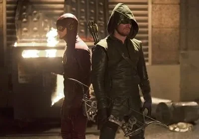 Dois heróis mascarados, um de vermelho com raio e outro de verde com arco, de costas numa rua escura e urbana.