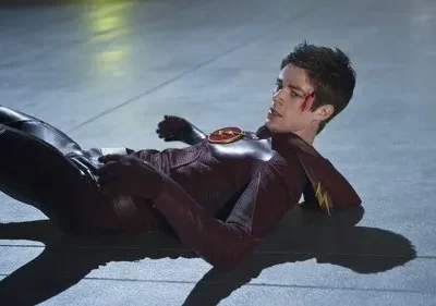 Homem em traje vermelho de super-herói, com emblema de raio, deitado e ferido num chão cinzento. Há um corte na testa e ele parece desorientado.