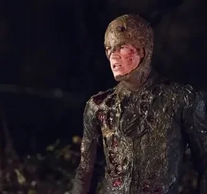 Homem com traje escuro e capuz, coberto de lama e ferimentos sangrentos, expressa angústia na escuridão de uma noite.