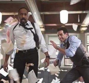 Dois detetives, um negro e um branco, reagem assustados em um escritório com papéis voando. Um usa coldre de ombro.