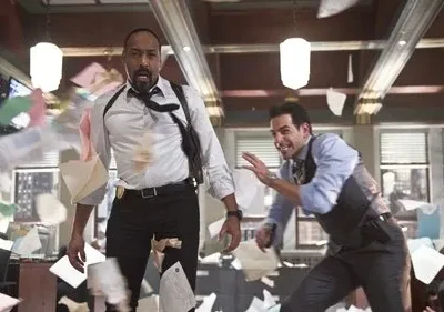 Dois detetives, um negro e um branco, reagem assustados em um escritório com papéis voando. Um usa coldre de ombro.